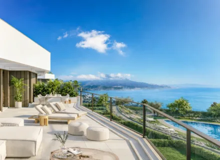 Spacious penthouse terrace with loungers and glass railing overlooking pool, coastline and mountains on a sunny day.