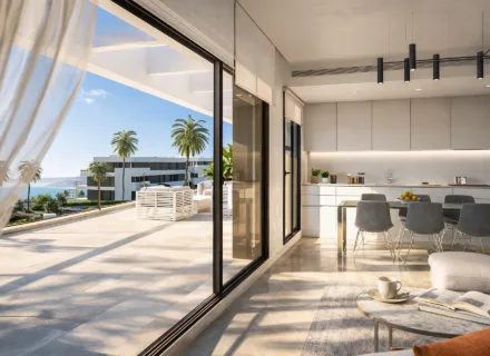 Bright modern penthouse interior with open kitchen and large terrace overlooking palm trees and the sea on a sunny day.