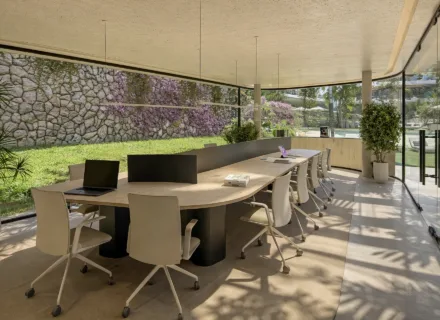 Contemporary co-working area with shared desks, white chairs and floor-to-ceiling windows overlooking lush garden.